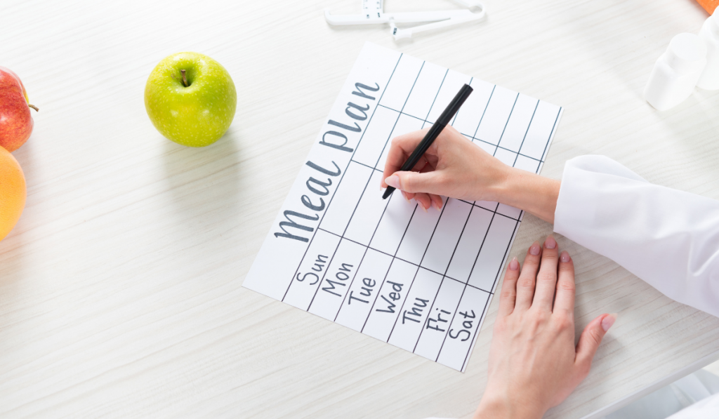 cropped view of dietitian writing in meal plan at table with fruits
