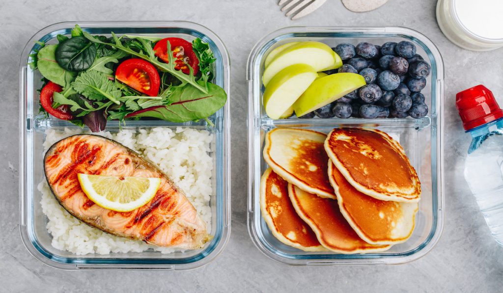 two meal prep containers with salmon and salad, pancakes and blueberries